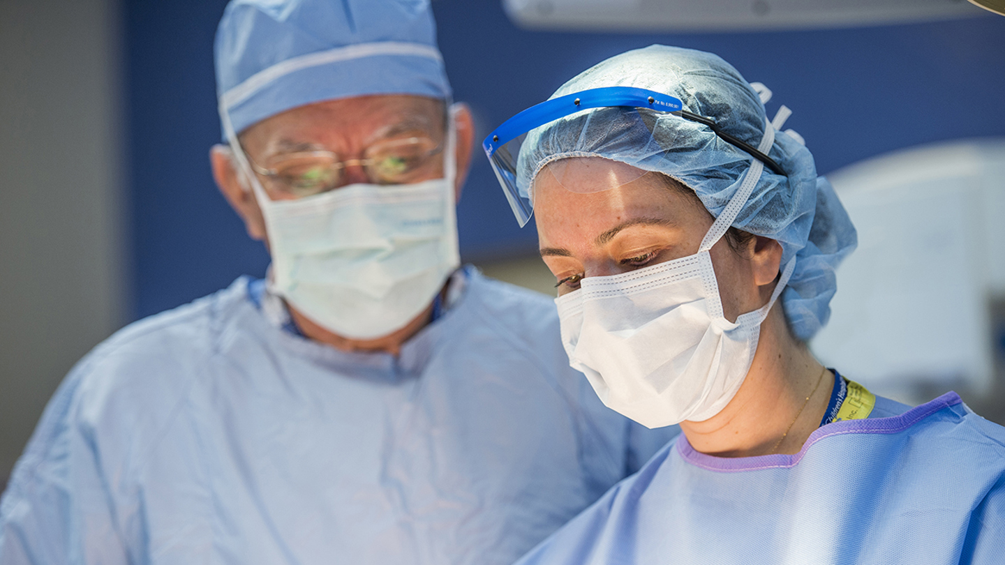 Alberto Peña, M.D. and Andrea Bischoff, M.D. are preforming colorectal surgery. Both surgeons are wearing masks and blue scrubs. 