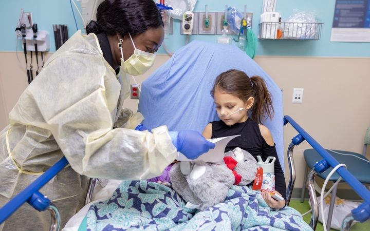 Woman wearing mask and PPE caring for a young girl holding a teddy bear sitting in a pediatric hospital bed. 