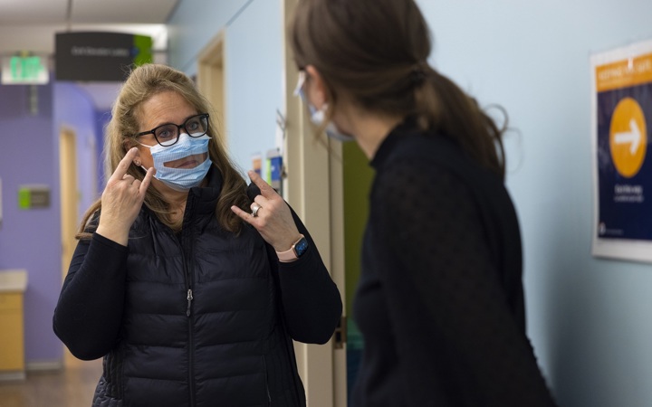 One woman using sign language and wearing a see-through mask, communicating with another woman. 