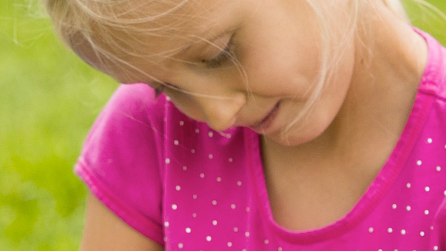 A girl in a pink shirt with sparkles sits in the grass, looking down at something and smiling.