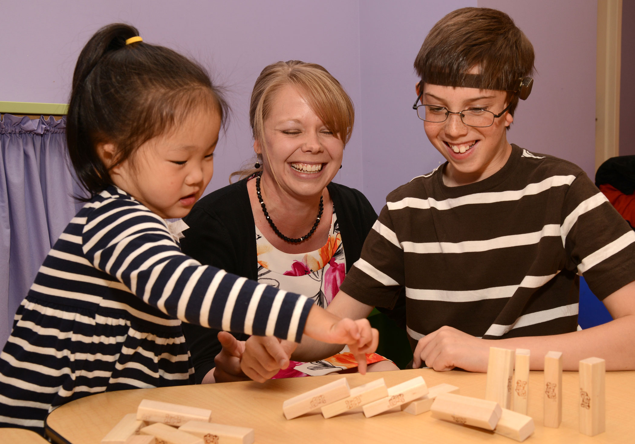 Audiologist Dr. Hedman sits in the middle of two patients playing Jenga. The girl on the left as her black hair in a ponytail and is wearing a black and white striped shirt. The boy on the right has glasses and is wearing a brown shirt with white stripes.