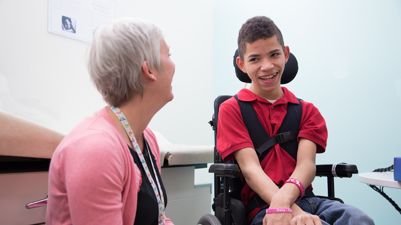 A boy wearing a red shirt sits in a wheelchair laughing with his doctor who is wearing a pink cardigan.