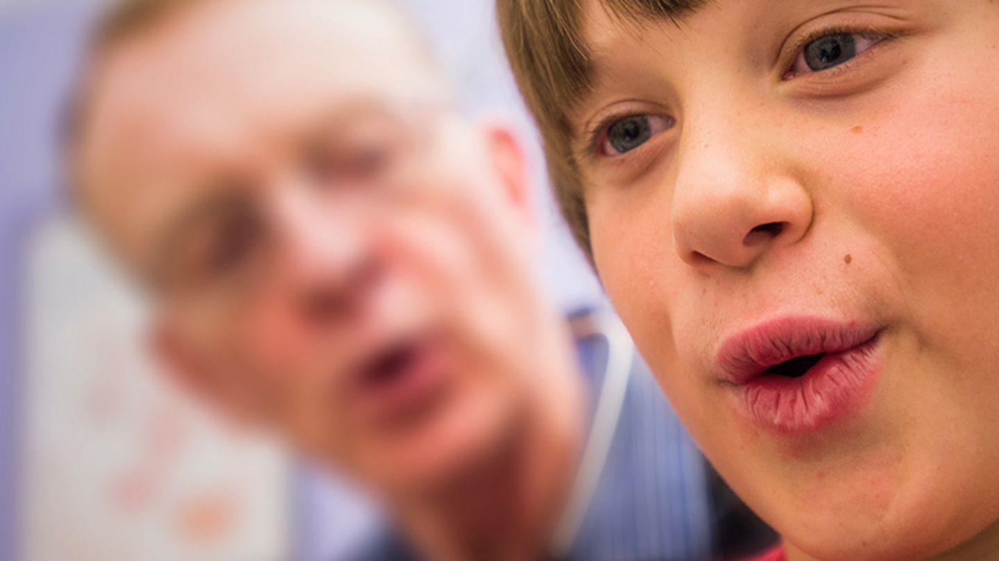 A boy with brown hair breathes out while his doctor watches in the background.