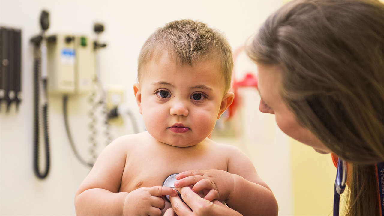 A woman with long brown hair uses a stethoscope to listen to an older baby's heart.