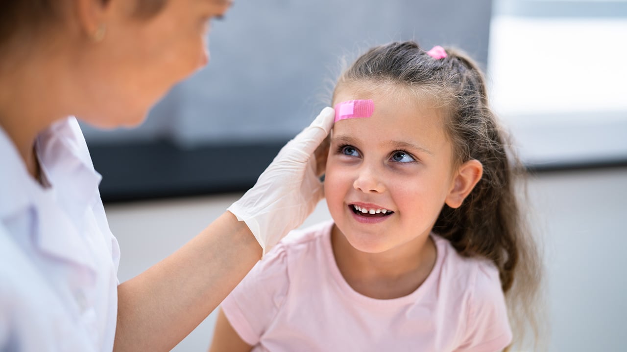 A medical professional places a pink band aid on the head of a small child in an office setting.  