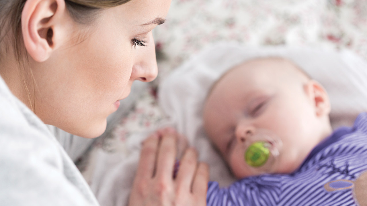 A woman with her eyes closed holds the hand of her sleeping baby.