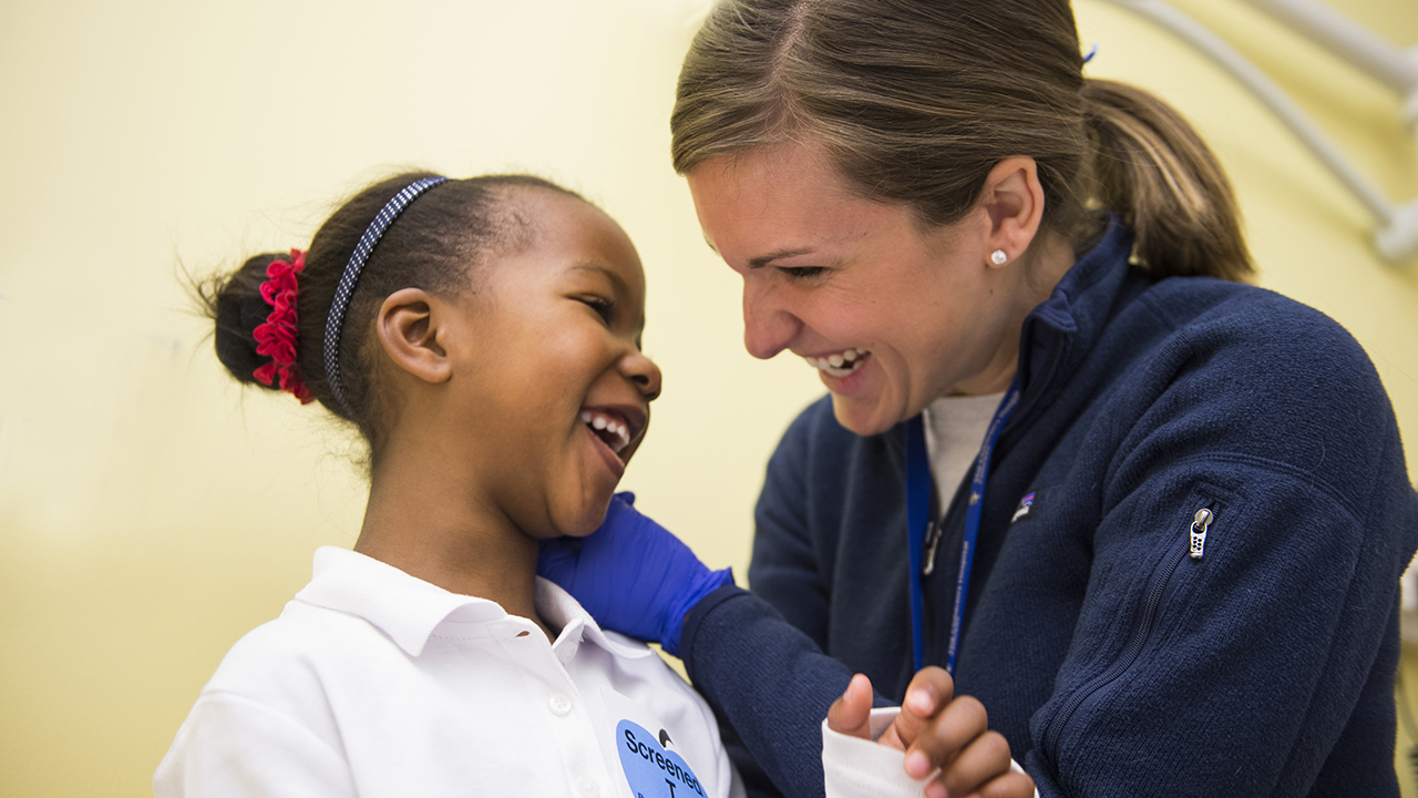 A woman with brown hair and wearing a blue fleece jacket smiles while examining a smiling girl's ear.