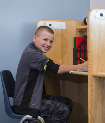 A boy with a buzz cut and wearing a gray and black zip up sweatshirt with black and gray camouflage pants sits at a wooden cubicle desk writing.